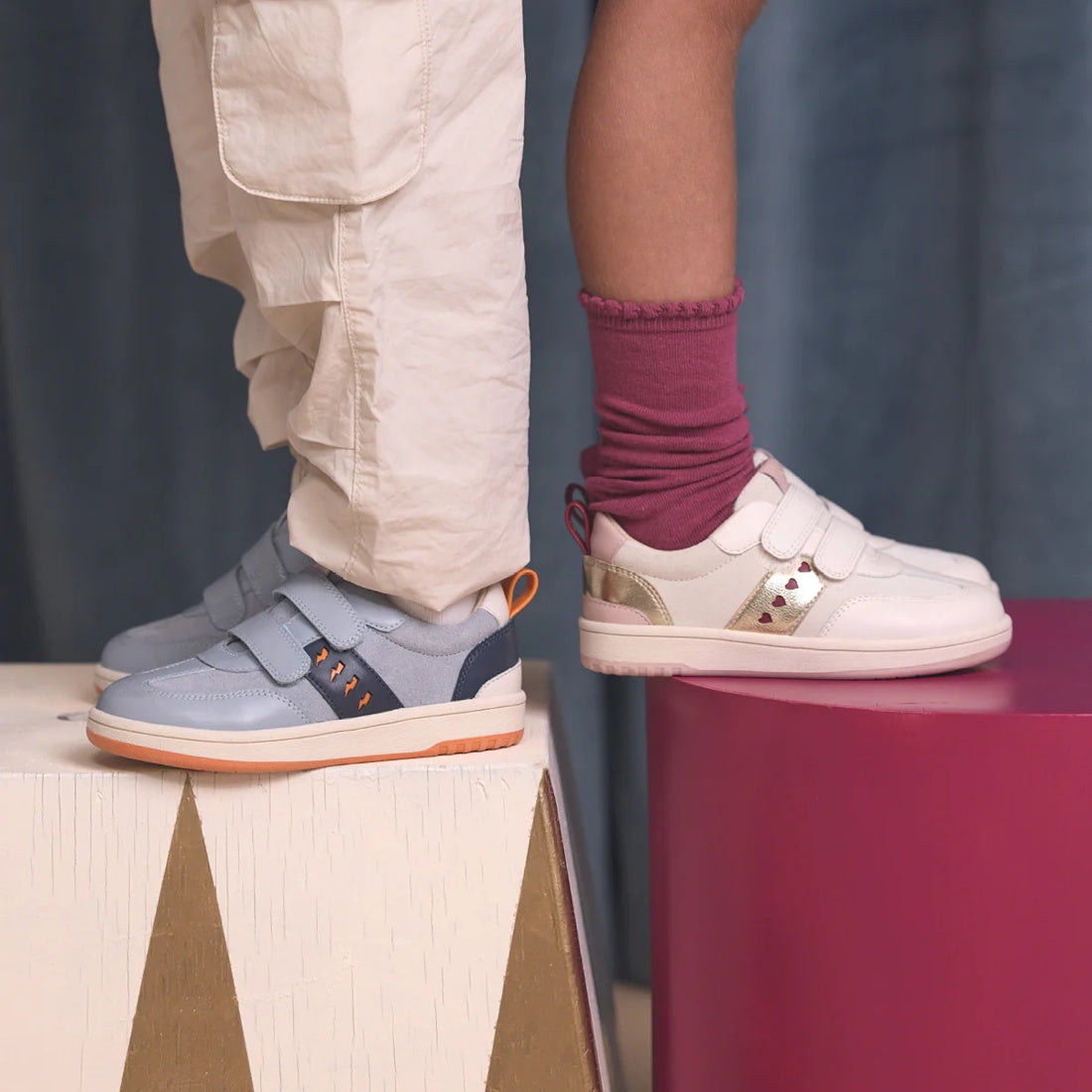 Two children stand on colorful blocks, showing only their legs and feet. One wears Pretty Brave River Bolts Leather Trainer Blue Grey with cream pants; the other wears white sporty shoes with pink socks and a skirt., 9420075363984, 9420075363977, 94200753