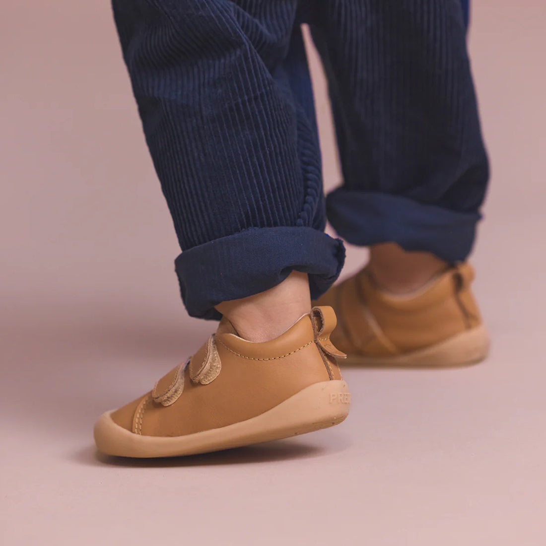 A child stands on a light pink surface, showing only their lower legs and feet dressed in rolled-up navy blue corduroy pants and Pretty Brave First Walker Toto Leather Trainer Tan shoes., 9420075366183, 9420075366176, 9420075366169, 9420075366152, 51694