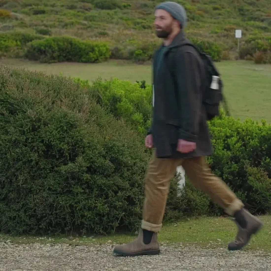 A man in a gray beanie, dark jacket, tan pants, and Blundstone 2446 Chelsea Boot - Vintage Clay Brown walks outdoors on a gravel path with green bushes and grass in the background., 9315891575034, 9315891575041, 9315891575058, 9315891575065, 9315891575072