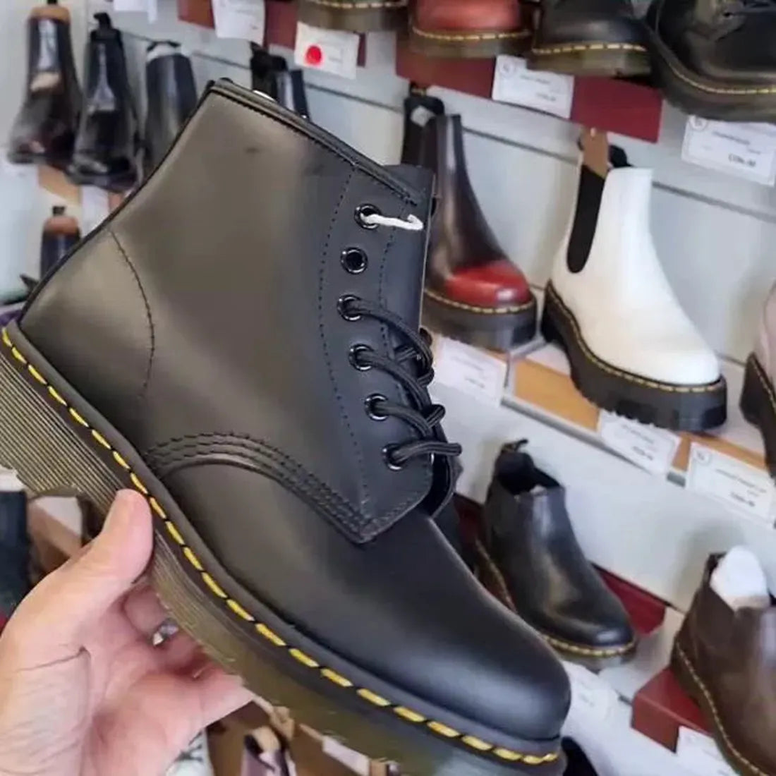 A hand shows the Dr Martens 101 6 Eye Yellow Stitching Boot, a sturdy black lace-up with signature sole, among colorful family shoes.  ksnz, 190665350715, 190665350722, 190665350739, 190665350746, 190665350753, 190665350760, 190665350777, 190665350784, 190665350791, 190665350807, 190665350814