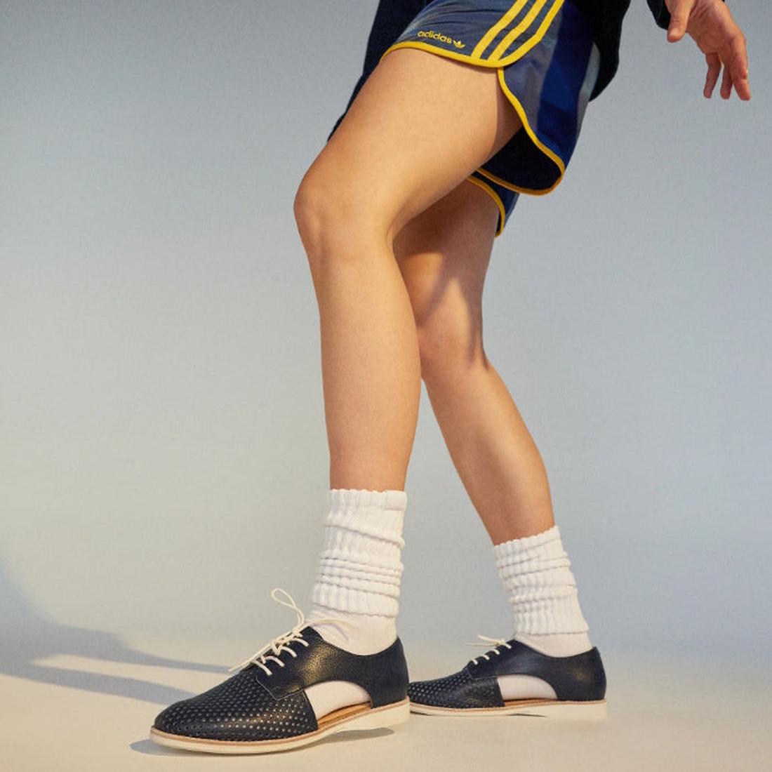 A person wearing navy and yellow athletic shorts, white socks, and Rollie Womens Sidecut Punch Navy Tumble Leather Shoes with anti-slip rubber outsoles and punch hole detailing steps forward against a neutral background., 9781760775414, 9781760775421, 978