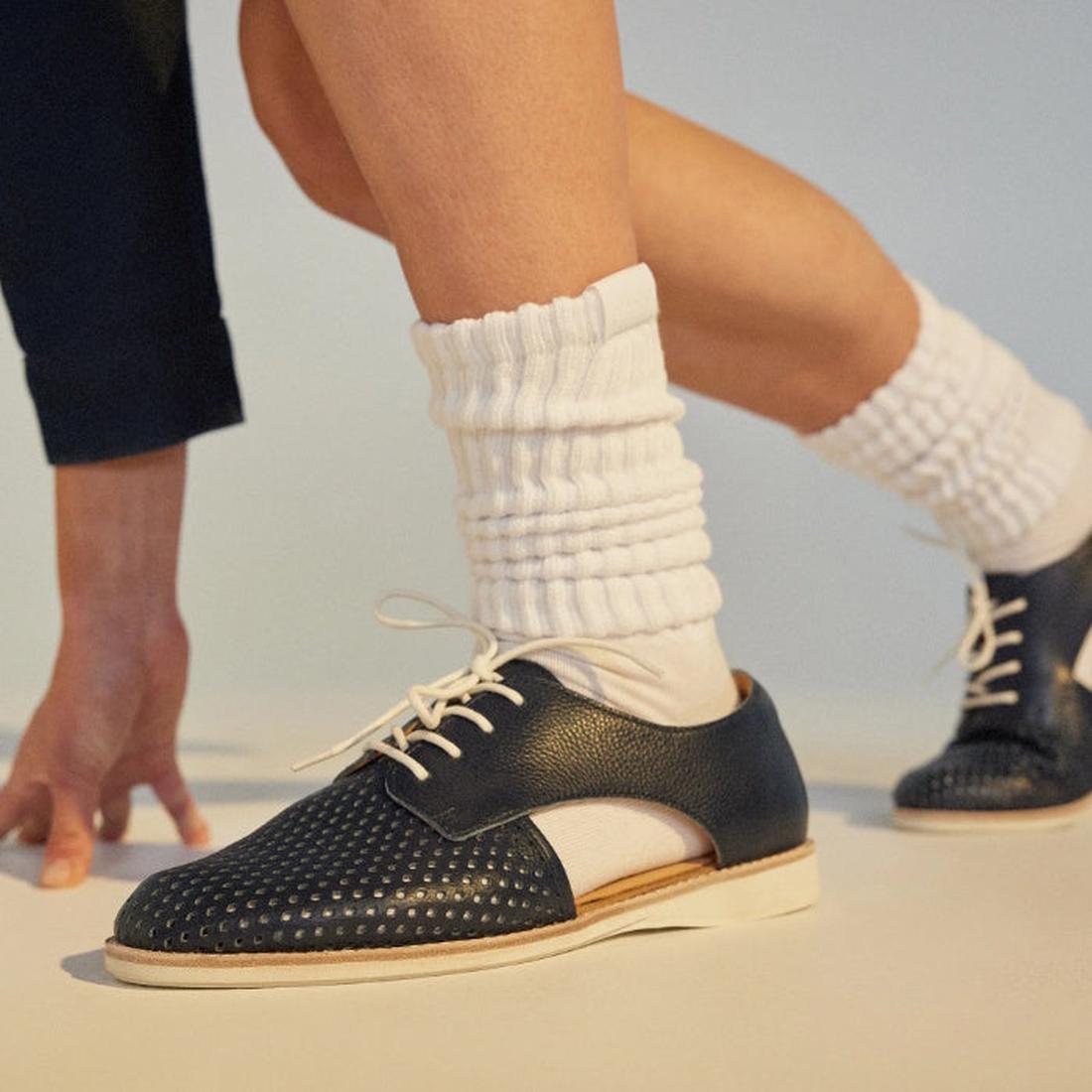 A person wears Rollie Womens Sidecut Punch Navy Tumble Leather Shoes with punch hole detailing and white laces, paired with thick white scrunched socks. One foot steps forward, ready to move—highlighting style and breathable linings., 9781760775414, 97817
