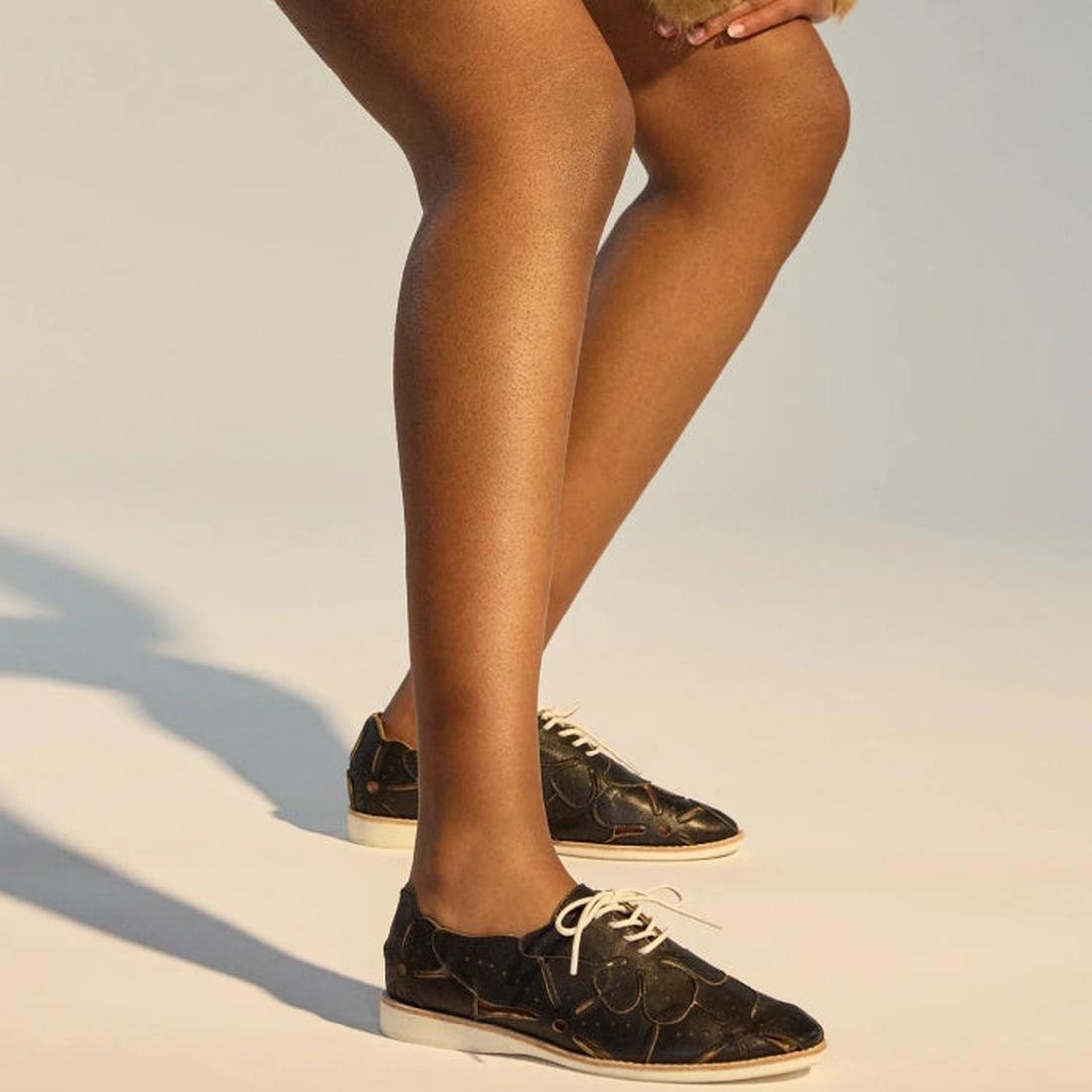From mid-thigh down, a person with brown skin is shown bending slightly, wearing the Rollie Derby Applique Black Womens Leather Brogue Shoe—Australian designed, featuring black patterned leather uppers and white laces., 9781760792312, 9781760792329, 97817
