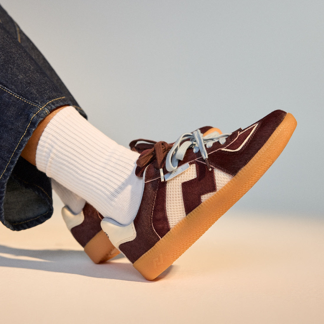 Wearing dark denim pants and white ribbed socks, a person rests one foot over the other, showcasing Rollie Womens Pace Dark Cherry Bone Suede Sneakers featuring a maroon and white suede upper, gum soles, and light blue laces., 9339554000315, 9339554000317