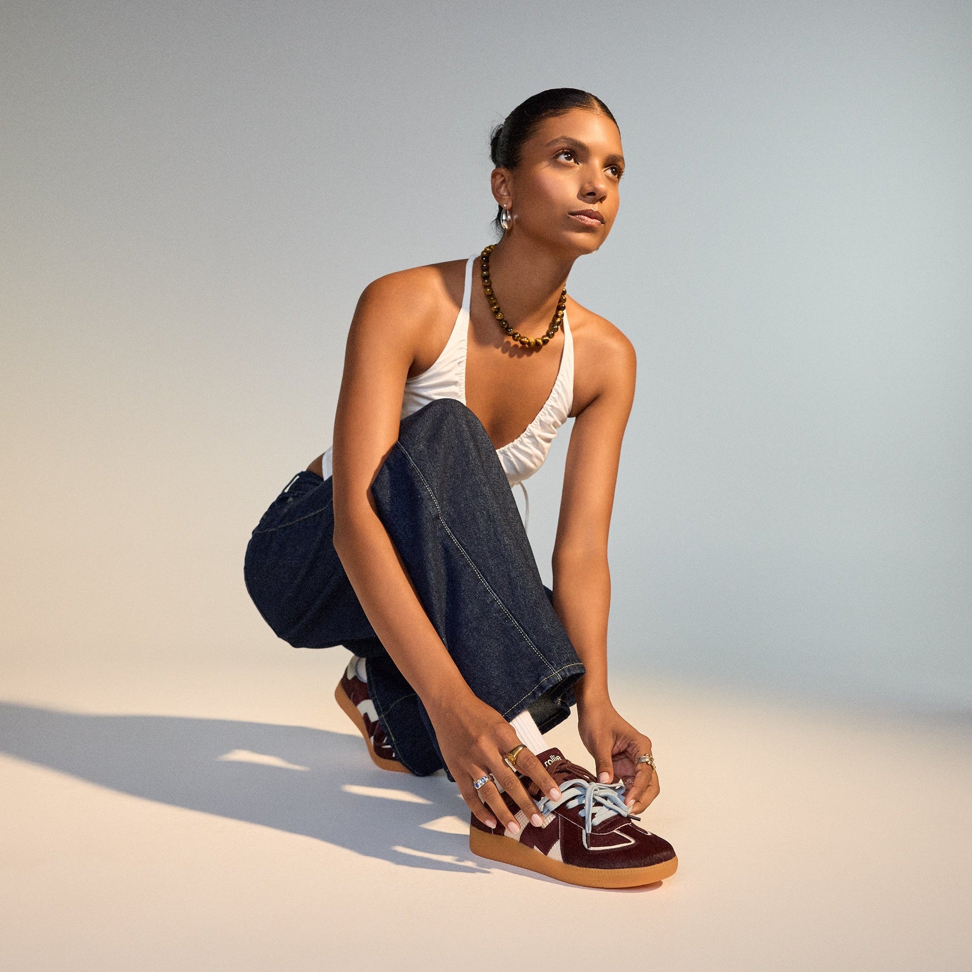 A woman crouches to tie her Rollie Womens Pace Dark Cherry Bone Suede Sneakers, styled with a white halter top, dark jeans, necklace, and rings, against a softly lit neutral background., 9339554000315, 9339554000317, 9339554000164, 9339554000056, 93395540