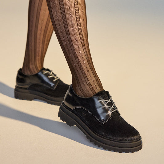 A person wears Rollie Womens Derby Step Black Splice Leather Brogues with comfy laces and a supportive sole, standing on pale flooring.  ksnz