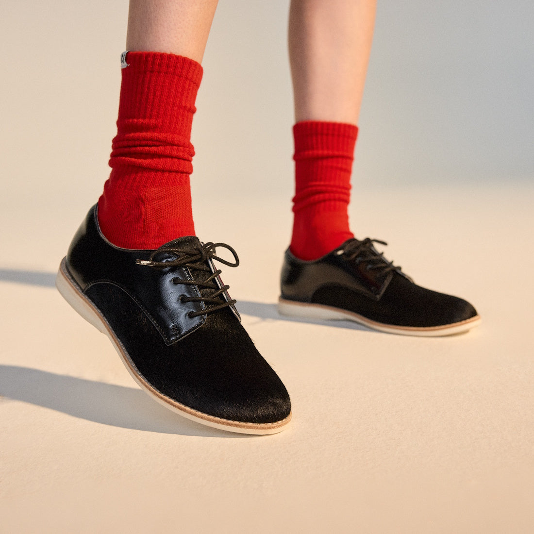 A person stands on a light surface wearing bright red socks and Rollie Womens Derby Black Splice Leather Brogue Shoes, featuring black lace-up design and anti-slip rubber outsoles. Only their lower legs and feet are visible., 51190, 9339554014920, 9339554