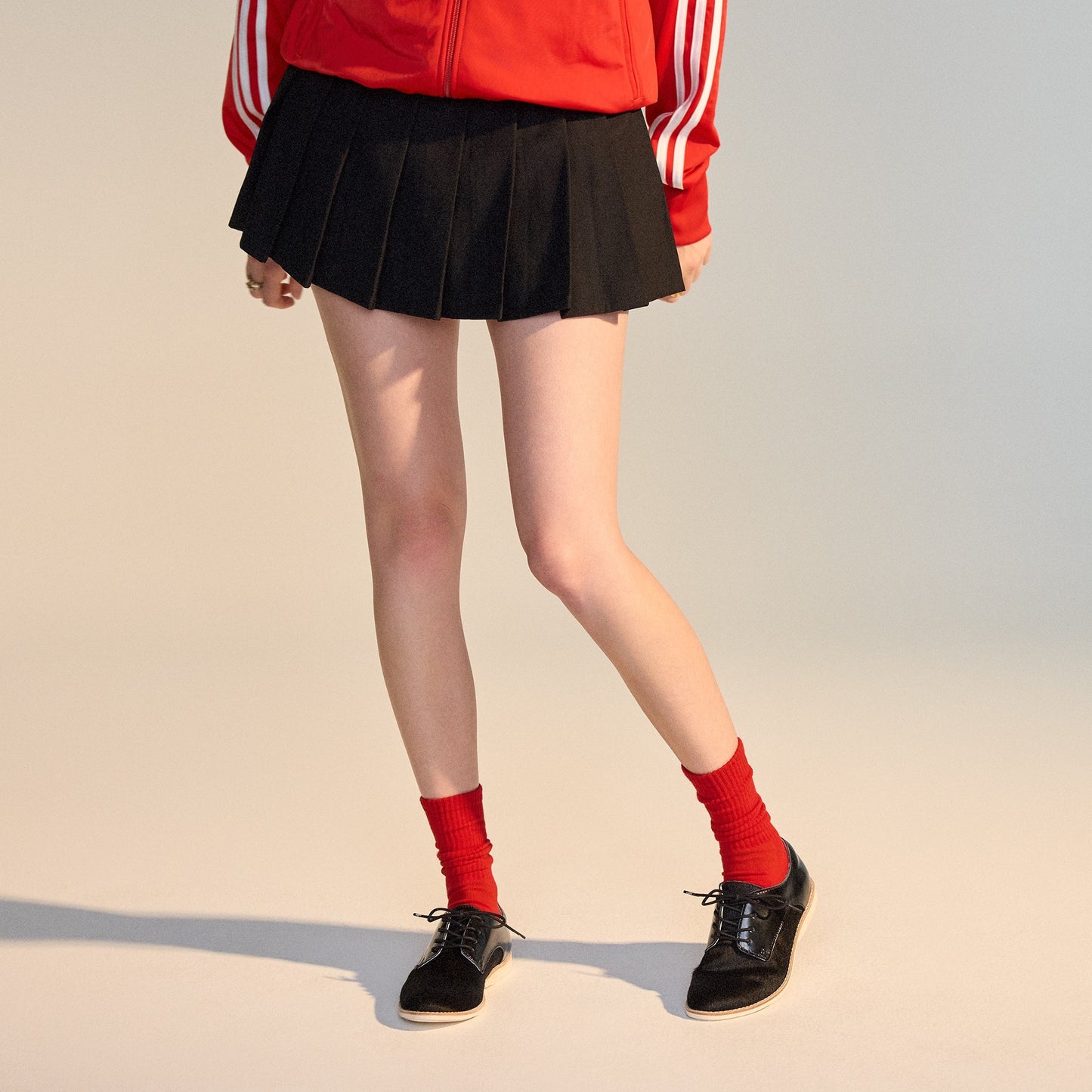 Person wearing a red jacket with white stripes, black pleated skirt, red socks, and Rollie Womens Derby Black Splice Leather Brogue Shoes with an anti-slip rubber outsole, standing against a plain background. Face not visible., 51190, 9339554014920, 93395
