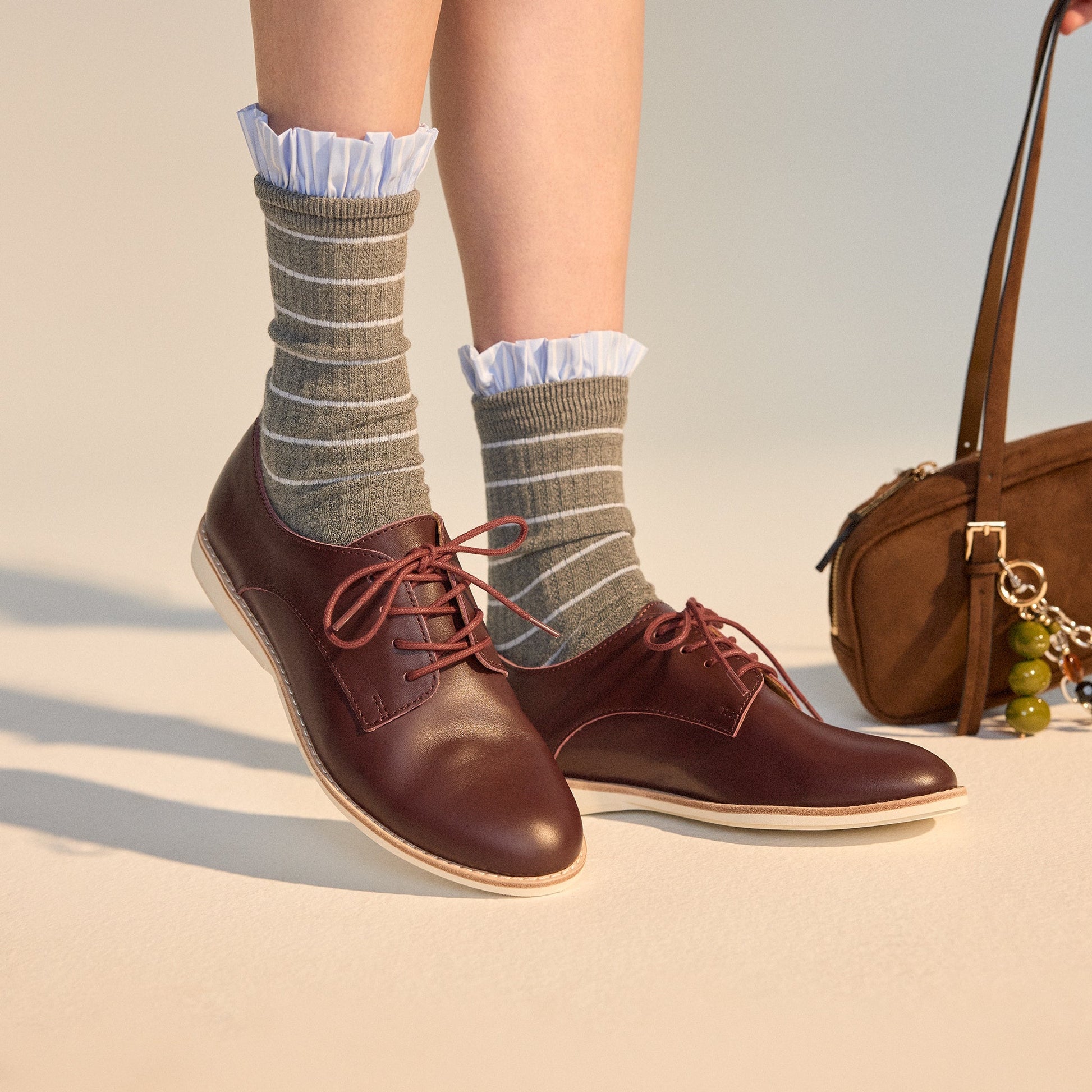 A person wearing gray striped socks with white ruffles and Rollie Womens Derby Super Soft Dark Cherry Leather Shoes stands on a light surface. A brown handbag with a green keychain is partially visible nearby., 9339554034843, 9339554086330, 9339554099231,