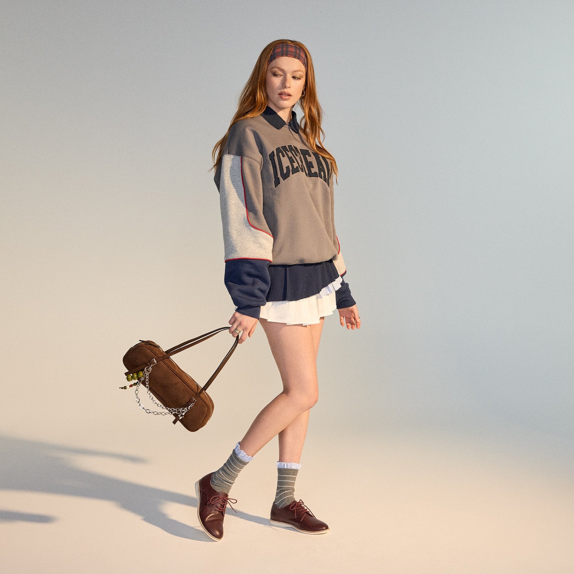 A young woman with long red hair wears a headband, an oversized ICECREAM sweatshirt, pleated white skirt, gray socks, and Rollie Womens Derby Super Soft Dark Cherry Leather Shoes. She holds a brown bag in a minimal, softly lit studio., 9339554034843, 9339
