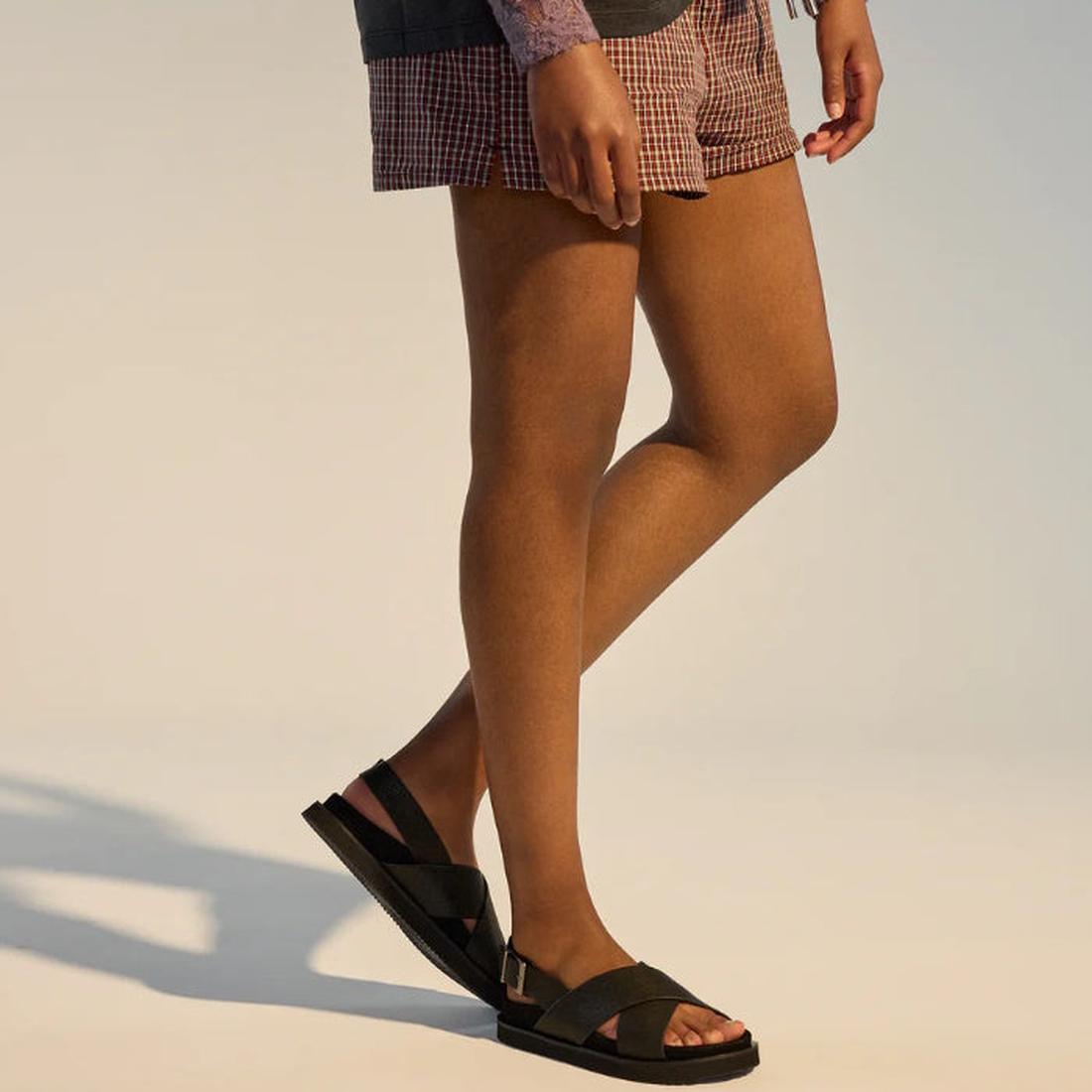 A person wears plaid shorts and Rollie Womens Rhodes Cross Sandal Tumble Black Leather with Back Strap, standing against a plain, light-colored background. Only the lower half of their body is shown., 48159, 9781760793692, 9781760793708, 9781760793715, 97