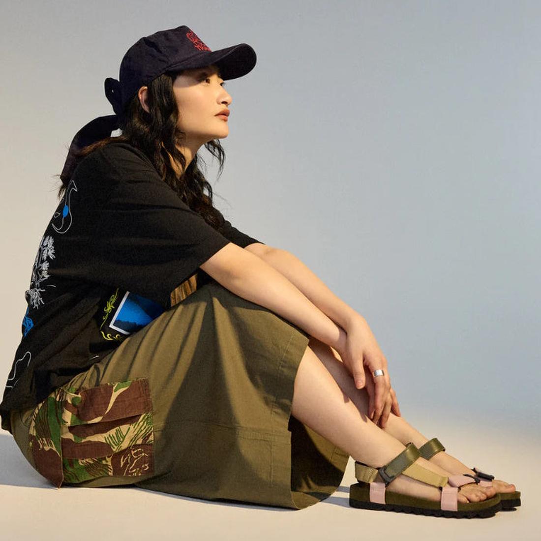 A woman sits sideways on the floor, wearing a navy cap, black tee with blue graphic, patchwork olive skirt, and Rollie Womens Sandal Tooth Wedge Blush Camo sandals with adjustable sheep leather straps. She looks ahead against a plain background., 93395543