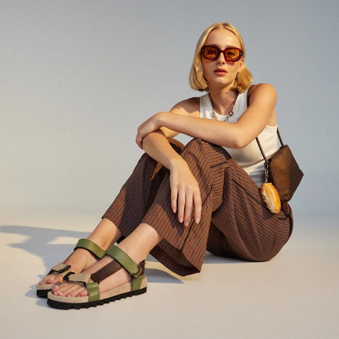 A woman with short blonde hair and sunglasses sits on the floor in a white sleeveless top, brown striped pants, and Rollie Womens Sandal Tooth Wedge Trail Green Brown, her black shoulder bag beside her and one arm resting on her knee., 48166, 978176079406