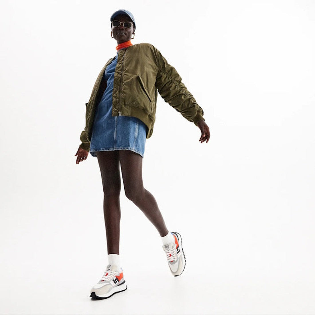 A person poses stylishly against a plain white background, wearing sunglasses, a blue cap, olive green bomber jacket, denim skirt, and Rollie Womens Kentu Royale Sneaker White Grey with orthotic-friendly support and anti-slip rubber outsole., 933955434385