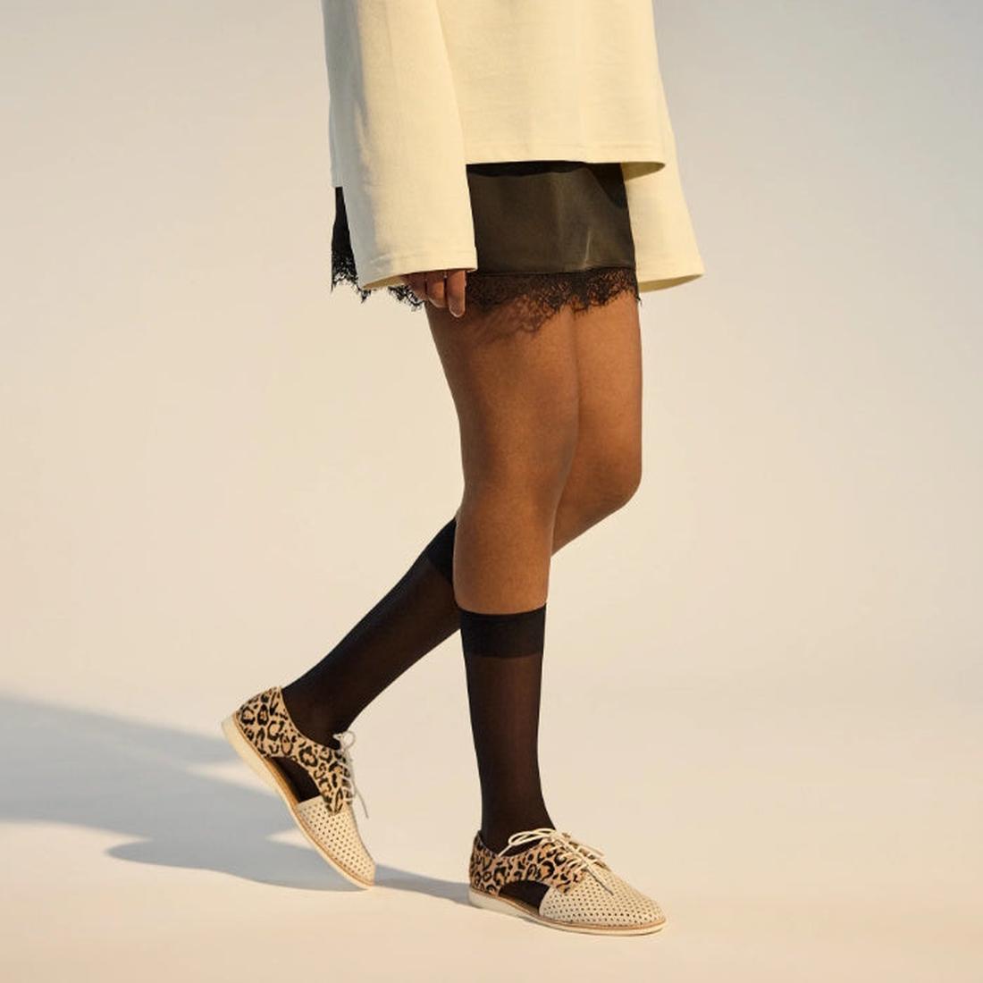 A person stands in a cream oversized top, black lace-trimmed skirt, sheer black knee-high socks, and Rollie Womens Sidecut Punch Bone Leather Leopard Cut Out Brogue shoes. The background is neutral., 9339554303413, 9339554303420, 9339554303437, 9339554303