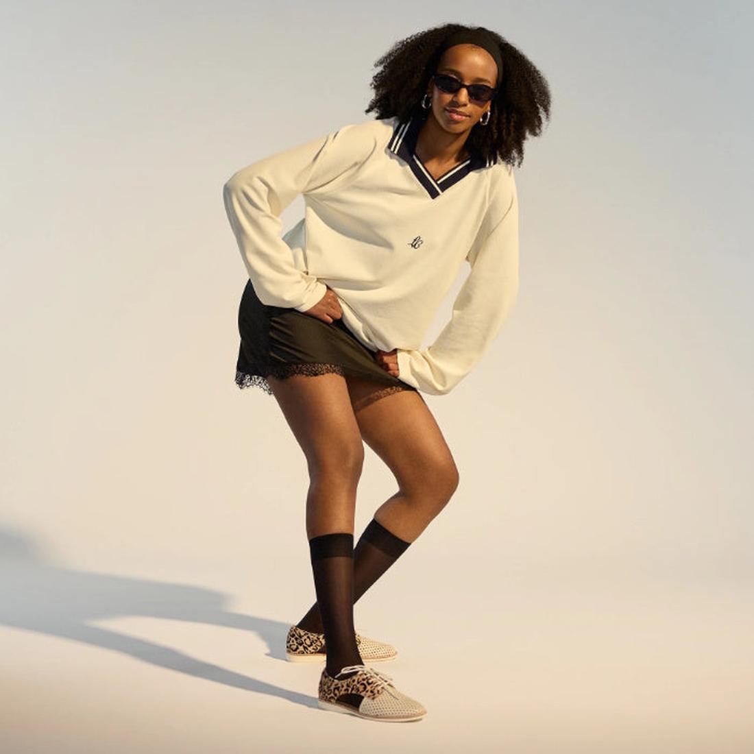 A woman with curly hair and sunglasses, in a cream sweater, black lace-trim mini skirt, black knee-high socks, and Rollie Womens Sidecut Punch Bone Leather Leopard Cut Out Brogue shoes poses against a plain background., 9339554303413, 9339554303420, 93395