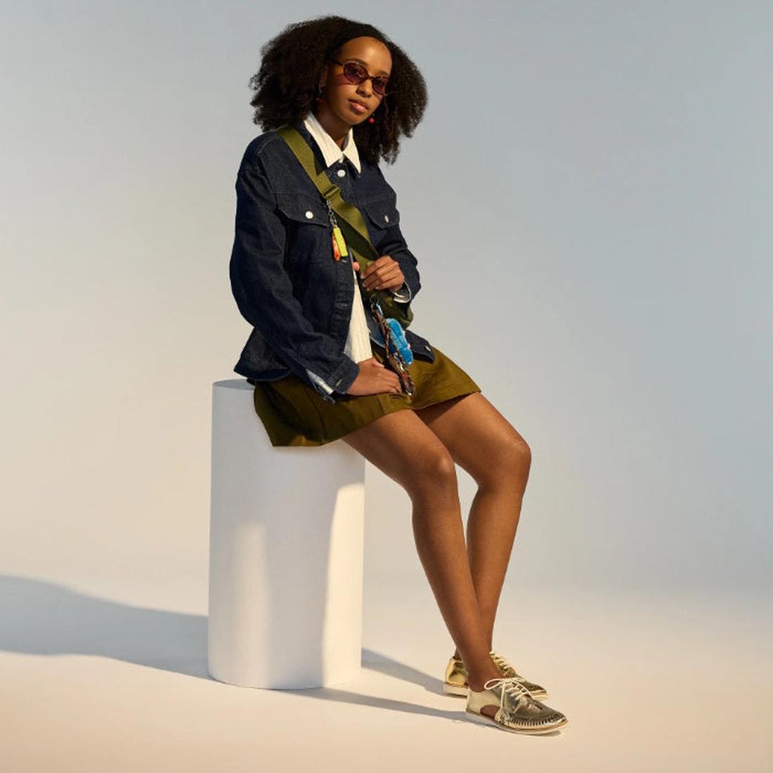 A woman with curly hair sits on a white stool wearing sunglasses, a denim jacket, white shirt, olive skirt, crossbody bag, and Rollie Womens Sidecut Boho Punch Gold Leather Shoes—all set against a neutral background., 9381760798811, 9381760798828, 9381760