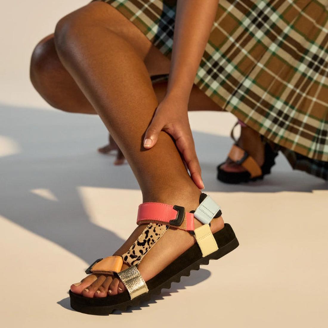 Kneeling, a person wears the Rollie Womens Sandal Tooth Wedge Leopard Multi—colorful wedge sandals featuring a suede footbed and adjustable metallic pink, orange, blue, and leopard-print leather straps on a black sole., 9339554333786, 9339554333793, 93395