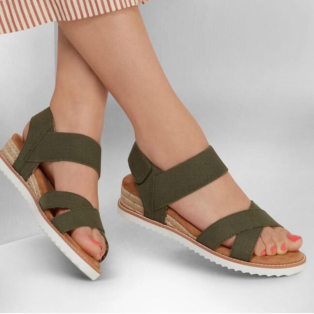 A person wears Skechers Bobs Womens Desert Kiss Secret Picnic Wedge Sandal in olive, featuring crisscross straps and a white ridged sole, paired with a striped skirt against a neutral background., 194428807249, 194428807256, 194428807263, 194428807270, 19
