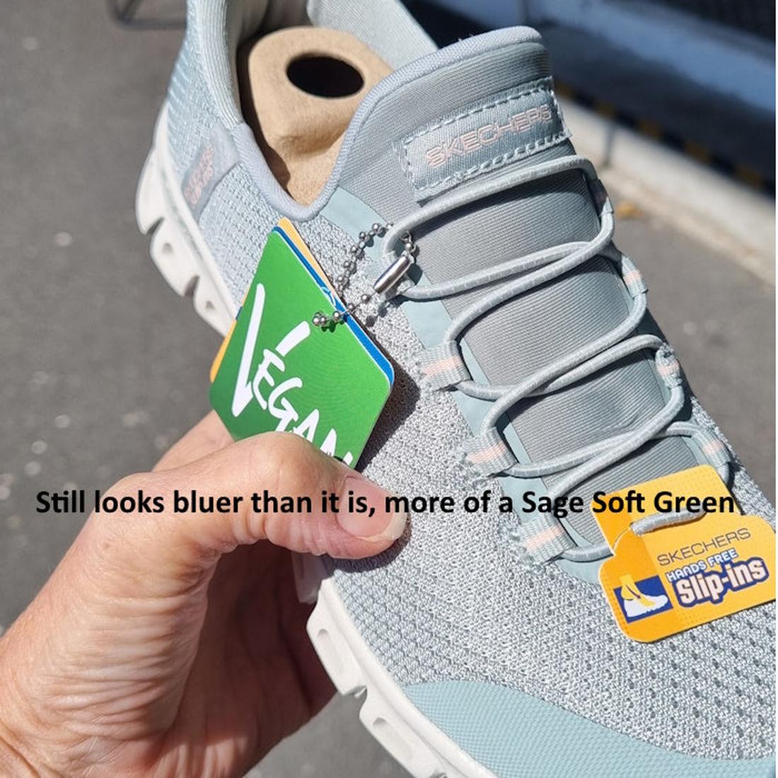 A hand holds a Skechers Womens Slip-Ins Glide Step Pursuit Sage sneaker labeled Vegan in sunlight. The shoe, with Air-Cooled Memory Foam, appears light blue but is called Sage Soft Green. Text overlays near the sneaker., 199025100960, 199025100984, 48989,
