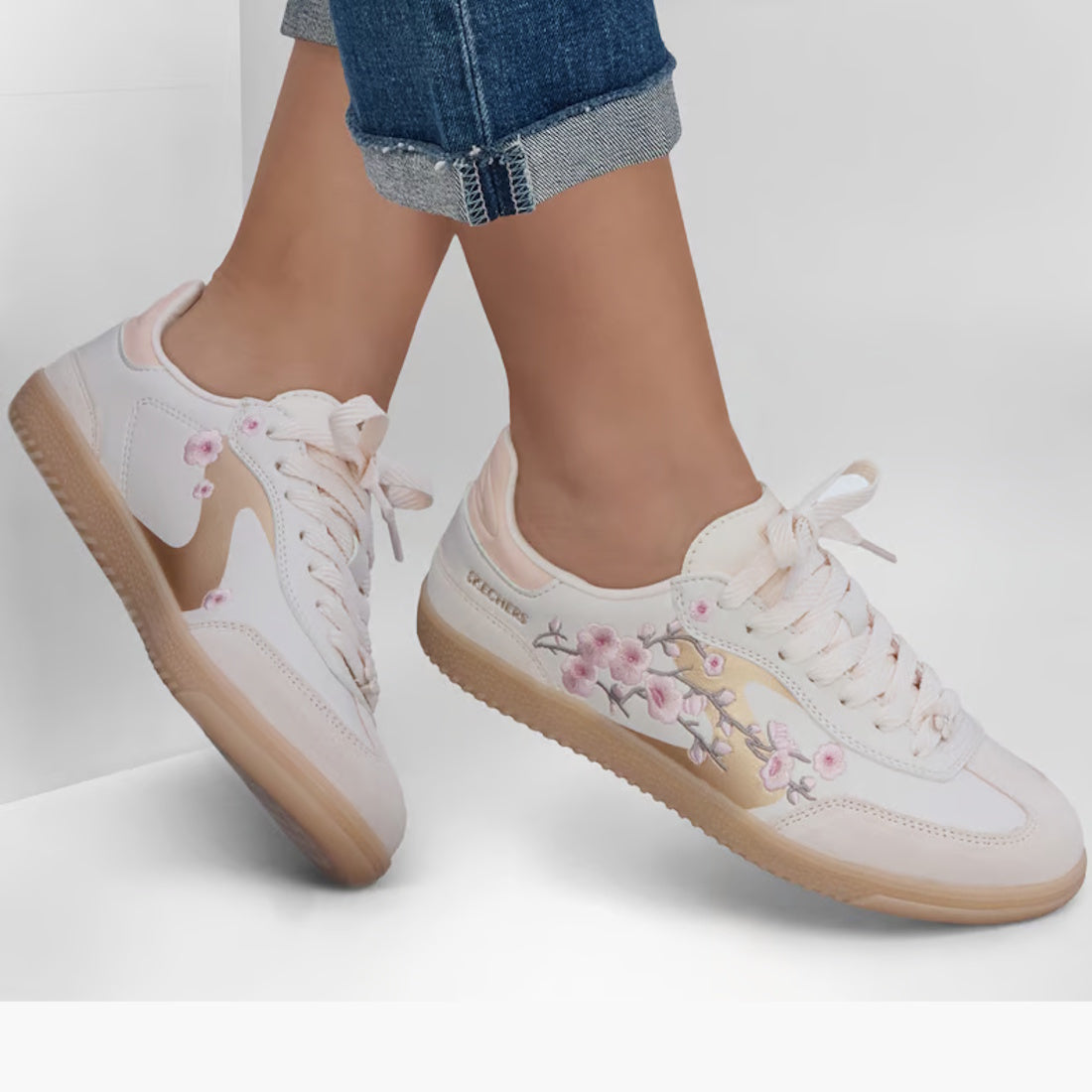 A person wearing rolled-up jeans and Skechers Womens HotShot Blooming Blossoms Court Shoe in light pink, adorned with floral embroidery and featuring Air-Cooled Memory Foam, stands on a white surface., 52721, 199025574884, 199025574907, 199025574921, 1990