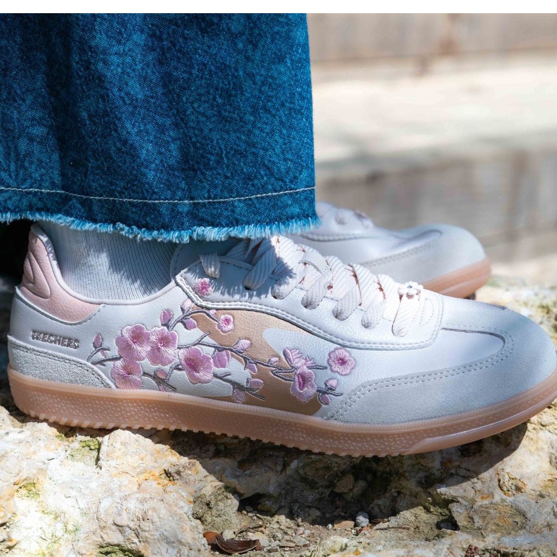 Frayed-hem blue jeans are paired with white floral sneakers featuring pink embroidery, such as the Skechers Women's HotShot Blooming Blossoms Court Shoe in Light Pink. The shoes rest on a textured stone surface in an outdoor setting., 52721, 199025574884,
