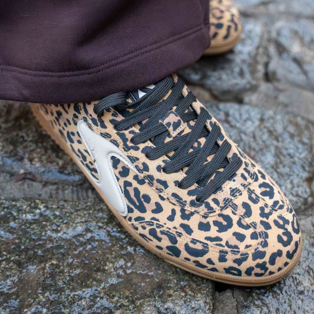 A close-up of someone wearing the Skechers Womens HotShot On The Prowl Leopard Suede Sneaker with brown and black leopard print, Air-Cooled Memory Foam, black laces, and white side detail, standing on stone pavement., 52252, 199025014359, 199025014373, 19