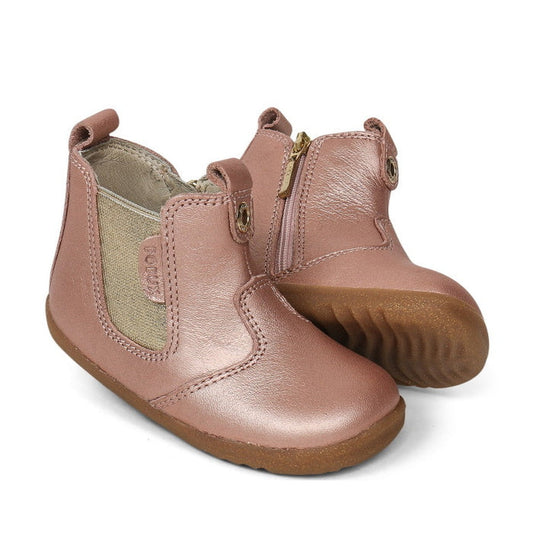 A pair of Bobux Step Up Jodphur Boot Rose Gold 721939 for children, featuring unisex design, rose gold leather, side zippers, elastic panels, and textured rubber soles. One boot stands upright while the other leans against it.