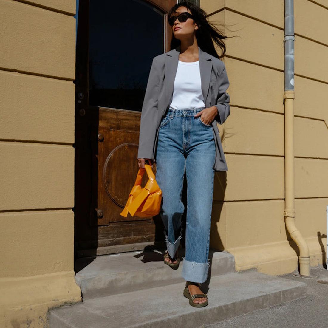 A woman in a gray blazer, white top, and wide-leg jeans walks confidently outside by a yellow wall. She carries a small orange bag, wears sunglasses, and completes her look with Woden Line Women's 3 Strap Sandal Dark Olive., 5713326432305, 5713326432312, 