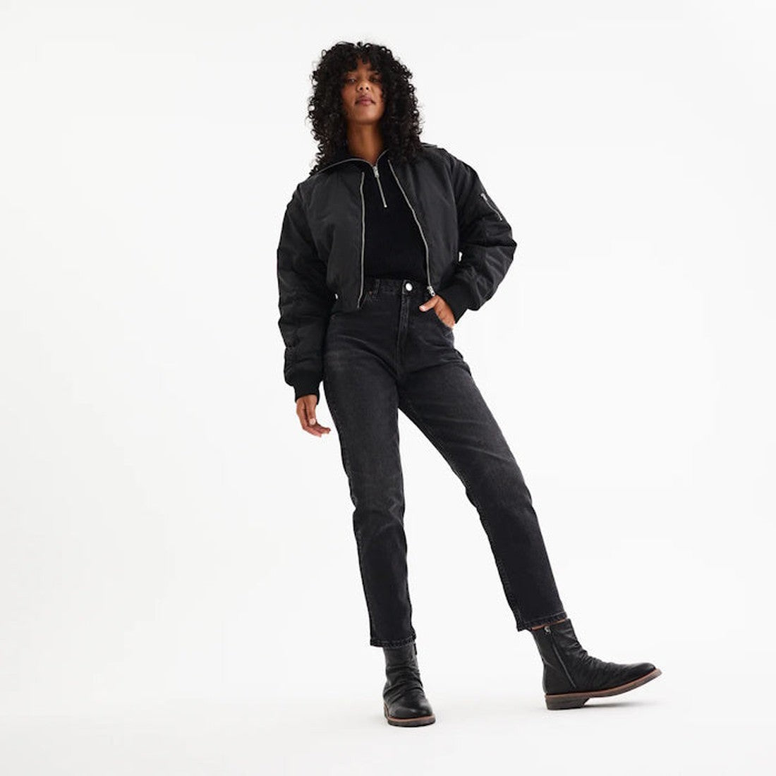 A person with curly hair wears a black bomber jacket, black top, high-waisted black jeans, and Rollie Womens Ankle Black Leather Boot Side Zip Rise Artisan, standing on a plain white background with one leg slightly bent., 9339554339214, 9339554339221, 93