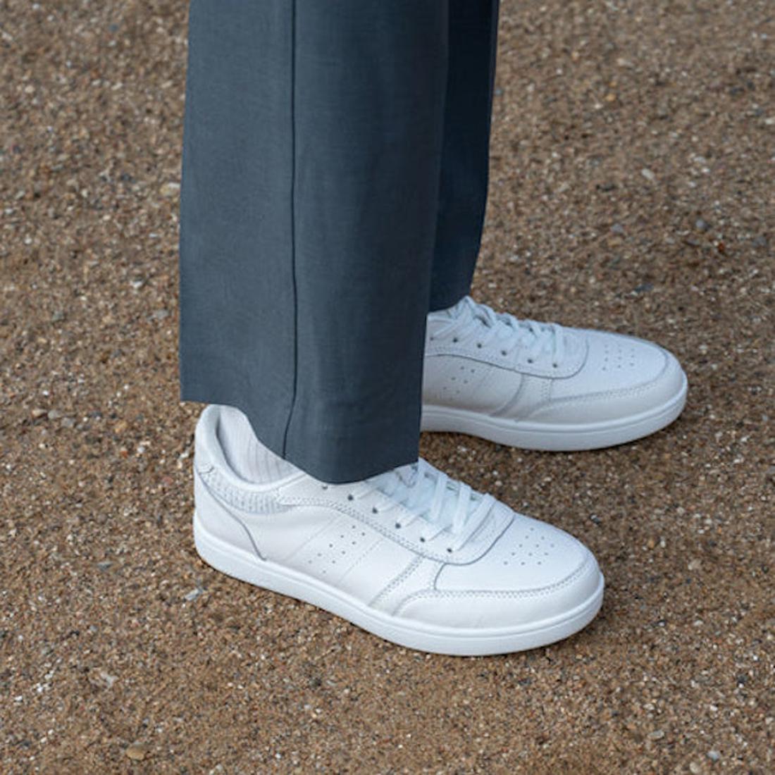 A person wearing dark gray pants and Woden Bjork Women's Leather Sneaker Blanc de Blanc White stands on gravel. The image is cropped to show only their lower legs and feet., 5713326460865, 5713326460872, 5713326460889, 5713326460896, 5713326460902