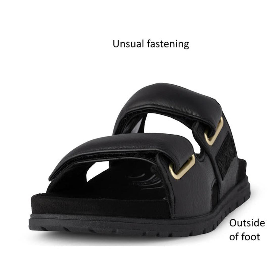 The Woden Lisa Women's Leather 2 Strap Sandal in black is shown from the front, featuring a unique fastening design, chunky buckle straps, and gold hardware. Text labels point out the distinctive fastening and outside of the foot., 5713326518276, 57133265