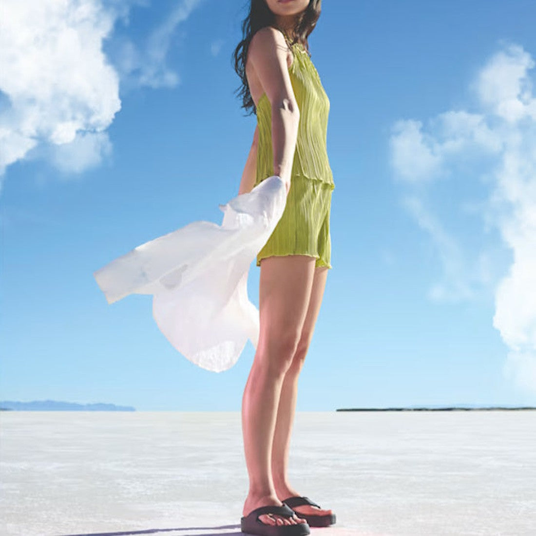 A woman in a sleeveless yellow romper stands on an open plain under a blue sky, holding a white jacket blowing in the wind. She wears Crocs - Womens Getaway PLATFORM Flip Sandal Black. Her face is partially out of frame., 42101, 196265540471, 196265540488